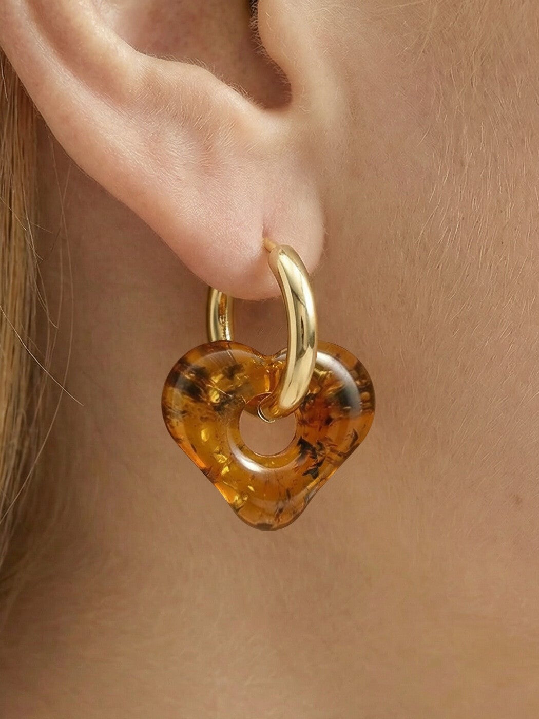 Nida Haven Amber Hoops, close-up of a soft heart-shaped charm of honey-brown amber on a gold-tone huggie hoop, worn on a woman's ear