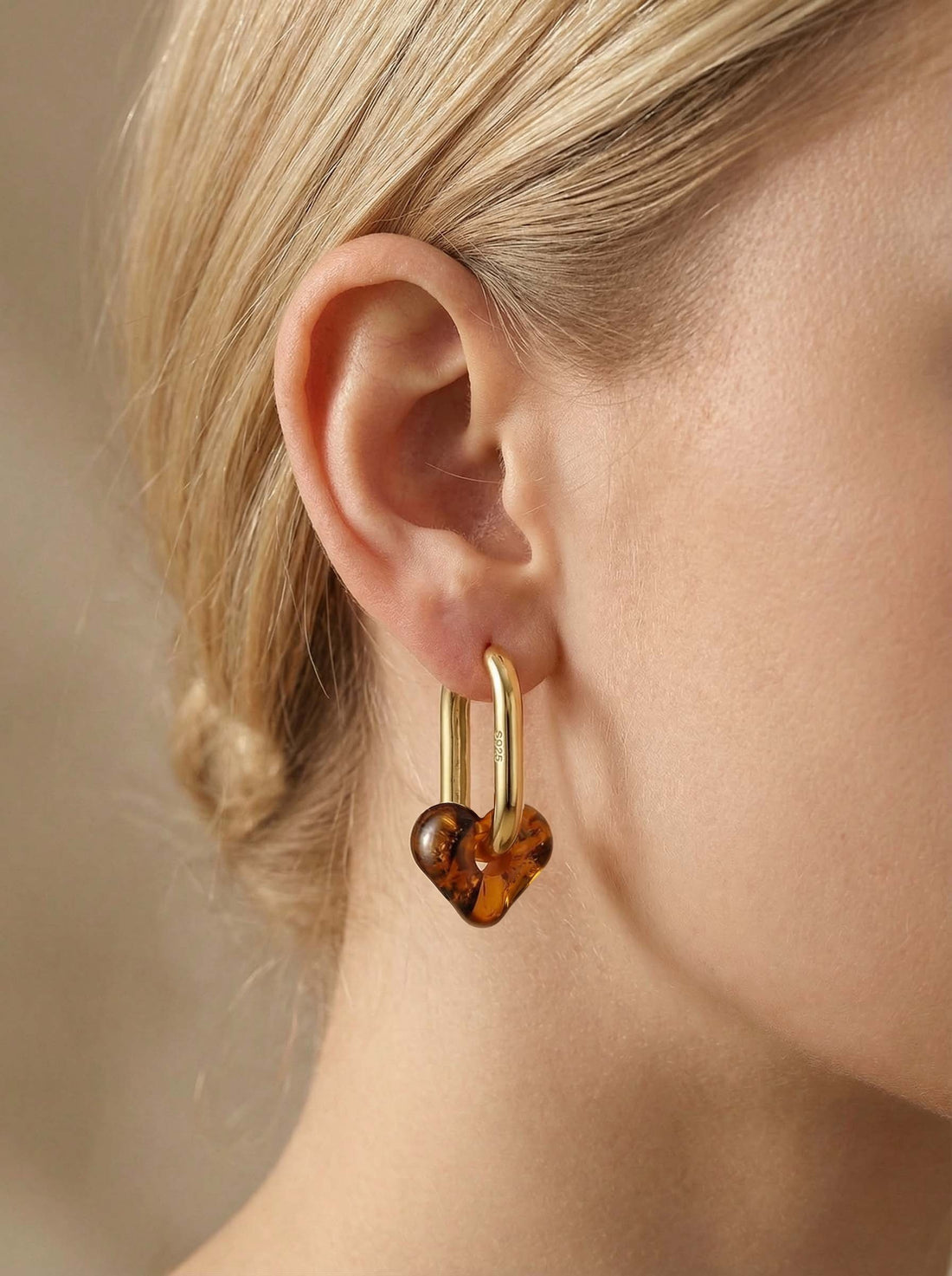 Dune Haven Amber Hoops, gold-tone tall oval hoop with a soft heart-shaped charm of honey-brown amber, worn on a woman's ear