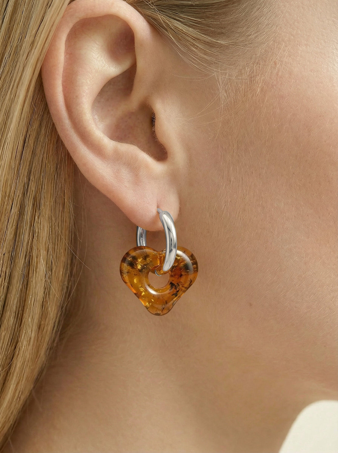 Nida Haven Amber Hoops, silver-tone huggie hoop with a soft heart-shaped charm of honey-brown amber, worn on a woman's ear