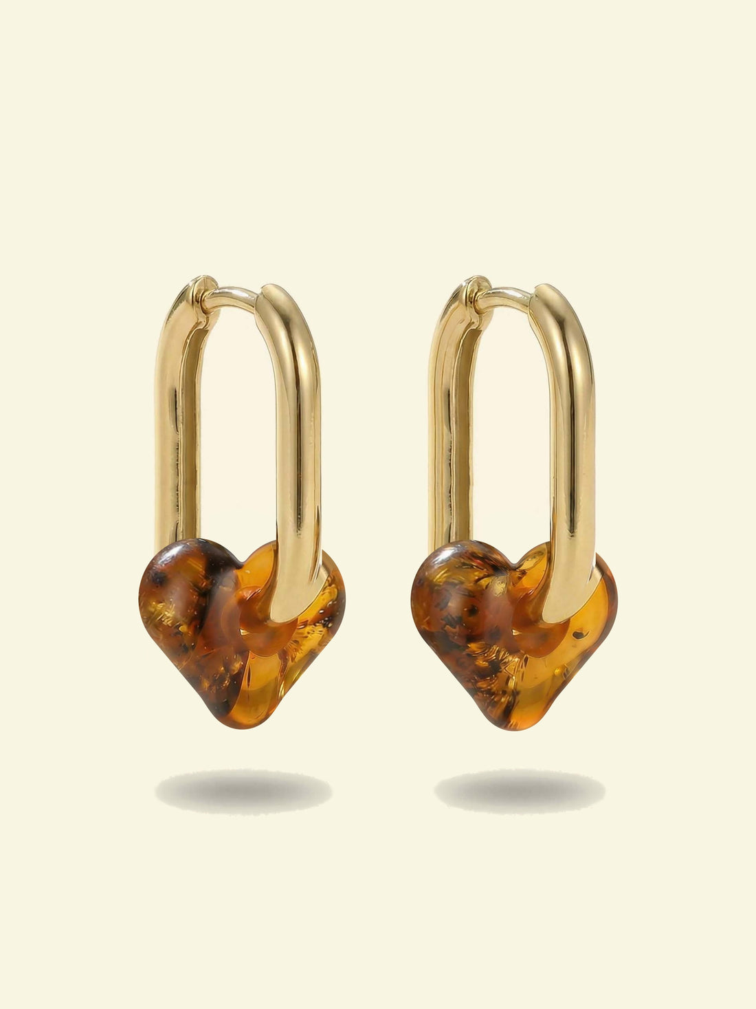 Dune Haven Amber Hoops, pair of gold-tone tall oval hoops with soft heart-shaped charms of honey-brown amber, front view on a cream background