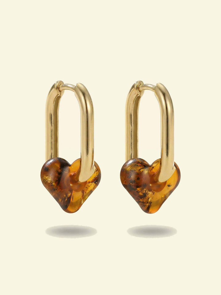 Dune Haven Amber Hoops, pair of gold-tone tall oval hoops with soft heart-shaped charms of honey-brown amber, front view on a cream background