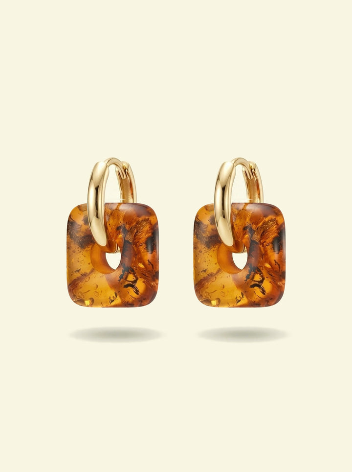 Nida Lantern Amber Hoops, pair of gold-tone huggie hoops with rounded square charms of honey-brown amber, front view on a cream background
