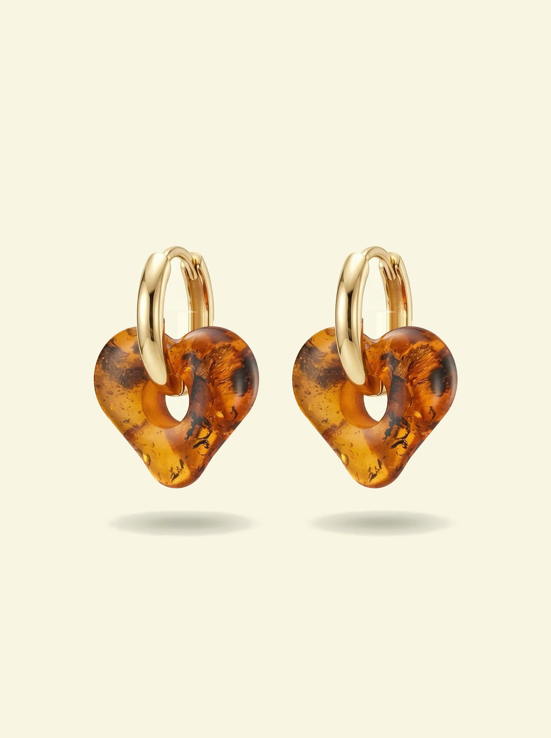 Nida Haven Amber Hoops, pair of gold-tone huggie hoops with soft heart-shaped charms of honey-brown amber, front view on a cream background
