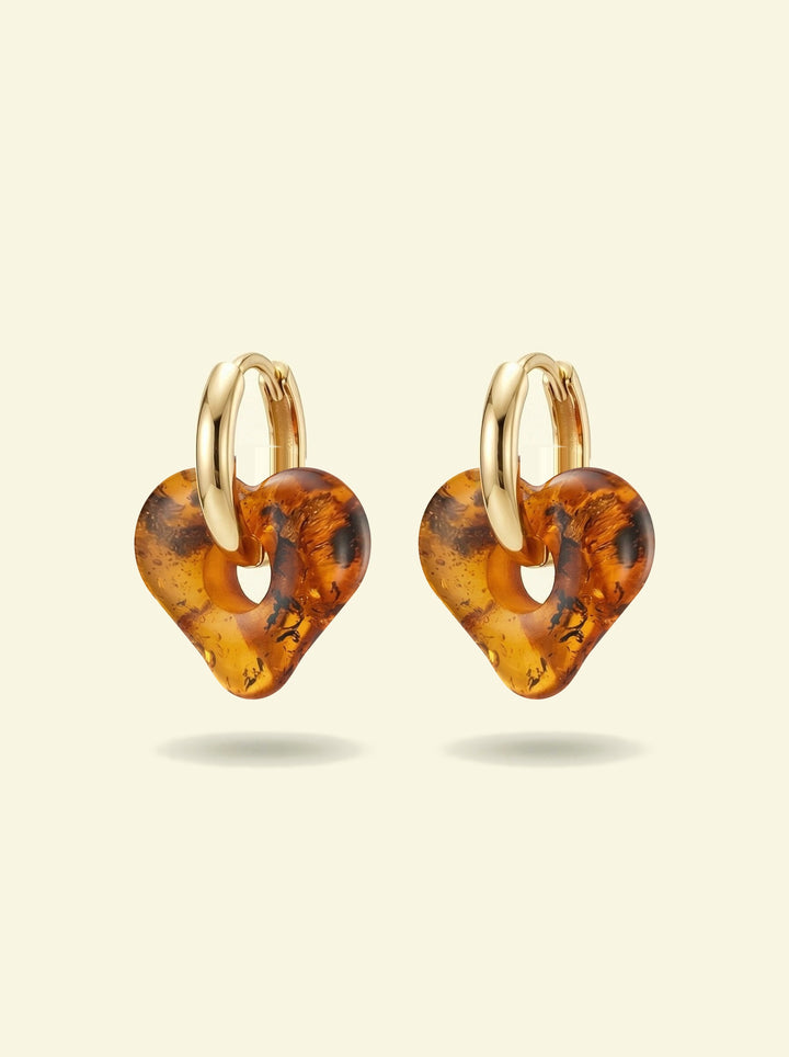 Nida Haven Amber Hoops, pair of gold-tone huggie hoops with soft heart-shaped charms of honey-brown amber, front view on a cream background