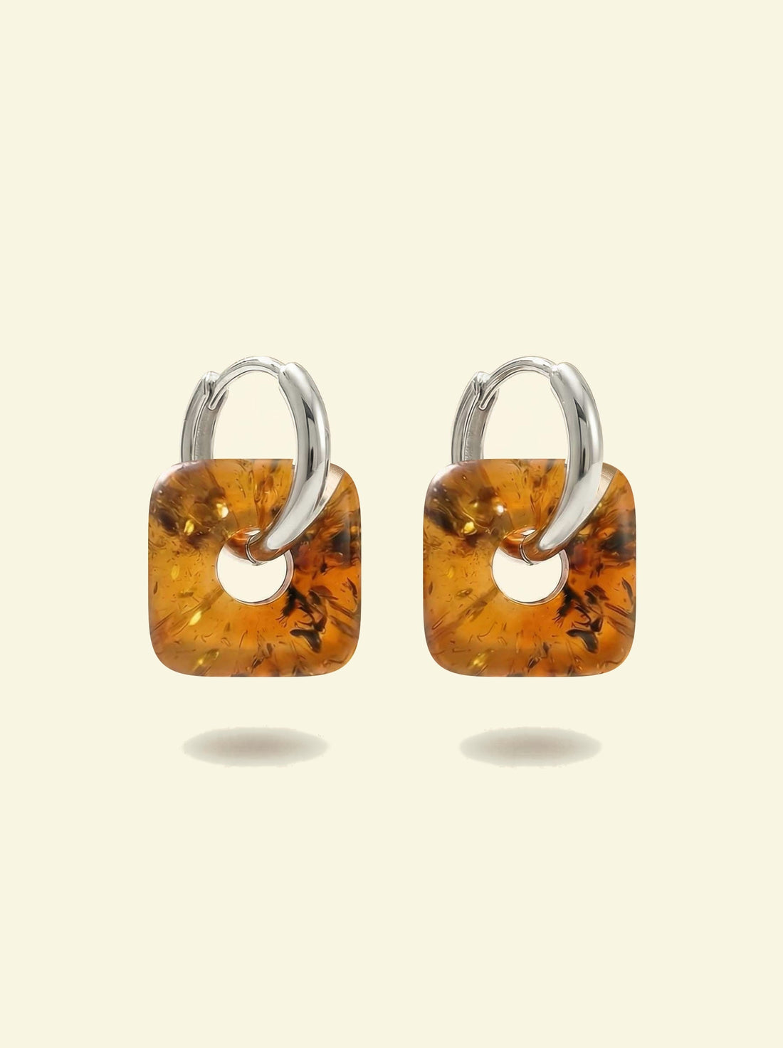 Nida Lantern Amber Hoops, pair of silver-tone huggie hoops with rounded square charms of honey-brown amber, front view on a cream background