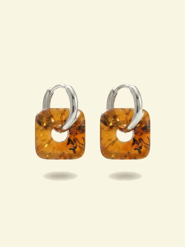 Nida Lantern Amber Hoops, pair of silver-tone huggie hoops with rounded square charms of honey-brown amber, front view on a cream background
