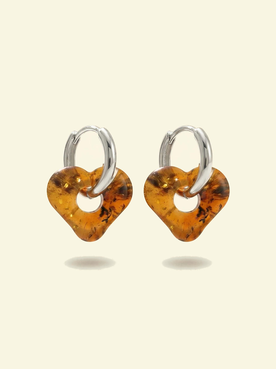 Nida Haven Amber Hoops, pair of silver-tone huggie hoops with soft heart-shaped charms of honey-brown amber, front view on a cream background