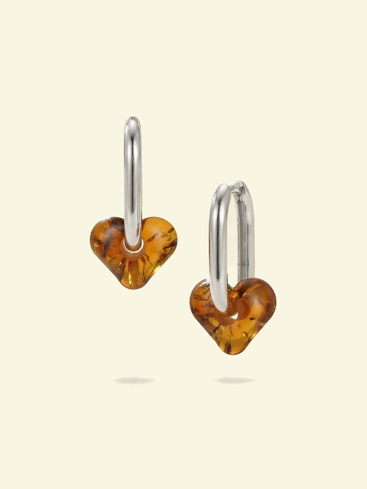 Dune Haven Amber Hoops, pair of silver-tone tall oval hoops with soft heart-shaped charms of honey-brown amber, front view on a cream background