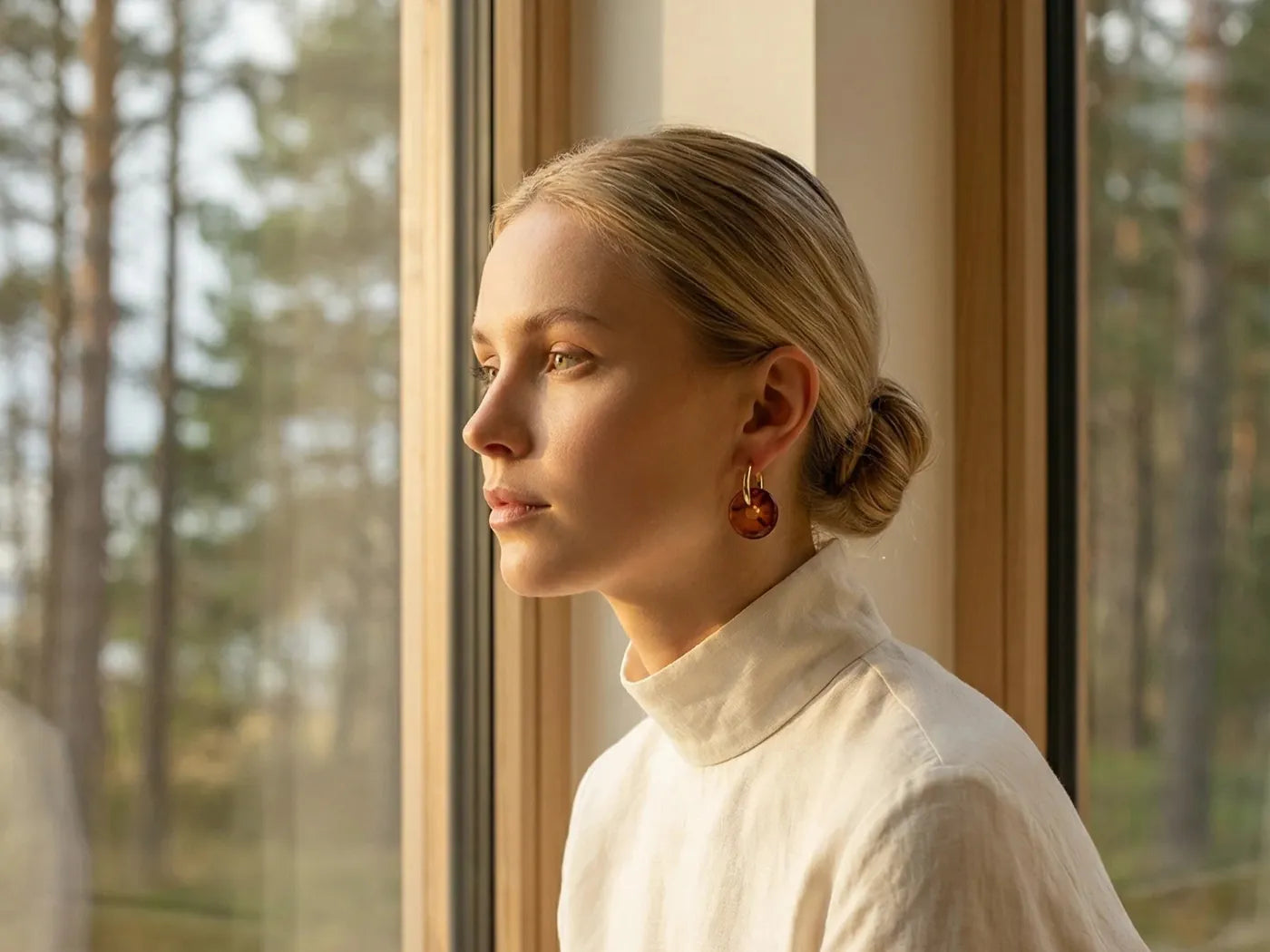 Woman in a cream turtleneck wearing amber earrings, looking out of a window onto a pine forest